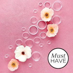 May include: Pink background with a variety of clear plastic circles of different sizes. Three pink flowers are arranged on the right side of the image. The text "Must HAVE" is in a black circle on the bottom right.