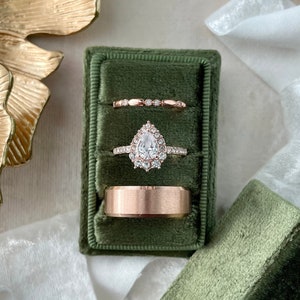 May include: Three gold rings in a green velvet ring box. The center ring features a pear-shaped diamond with a halo setting. The other two rings are plain bands.