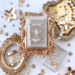 May include: A collection of gold rings with various gemstone settings, displayed in a gray velvet ring box. The rings are set against a backdrop of gold frames, pearls, and decorative floral accents, creating an elegant and luxurious presentation.