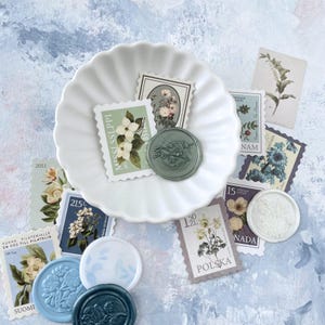 May include: A collection of vintage-style postage stamps and wax seals with floral designs. The stamps feature botanical illustrations and text, including "POLSKA" and "SUOMI." The seals are in shades of blue and green, arranged around a white, scalloped dish.