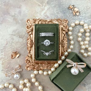 May include: An elegant display of jewelry featuring three rings in a green velvet box set within a gold ornate frame. A pearl necklace and a bow-shaped brooch with a pearl accent are also present, creating a luxurious aesthetic. The rings showcase sparkling gemstones.