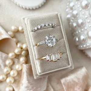 May include: Three engagement rings in a velvet ring box. The rings are all silver and have diamonds. The ring in the middle is a large oval-shaped diamond ring. The ring on the left is a thin band with small diamonds. The ring on the right is a band with five larger diamonds.