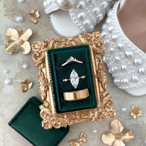 May include: A gold-framed ring box with a green velvet interior holds three rings: a gold band, a marquise-cut diamond ring, and a delicate crown ring. White pearl-studded bridal shoes and gold floral accents complete the wedding theme.
