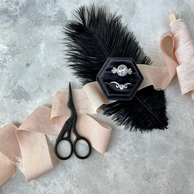 May include: A black velvet ring box with two silver rings, a black feather, and a pink satin ribbon. The box is on a gray background.