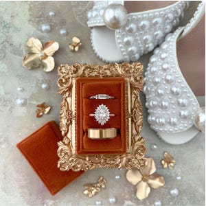 May include: A display of wedding rings in a decorative gold frame with a rust-colored velvet interior. The rings include a diamond halo engagement ring, a baguette diamond ring, and a gold wedding band. White pearl-studded bridal sandals and gold floral accents complete the arrangement.