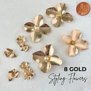 May include: Eight gold styling flowers in various sizes and designs. Some flowers have a pearl in the centre. The flowers are arranged on a white surface. A US penny is included for size reference.