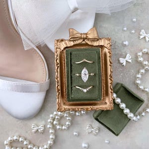 May include: A gold-framed ring box with three rings, including a marquise-cut diamond ring, sits on a surface with a white shoe, pearls, and decorative bows. The ring box has a green velvet interior.