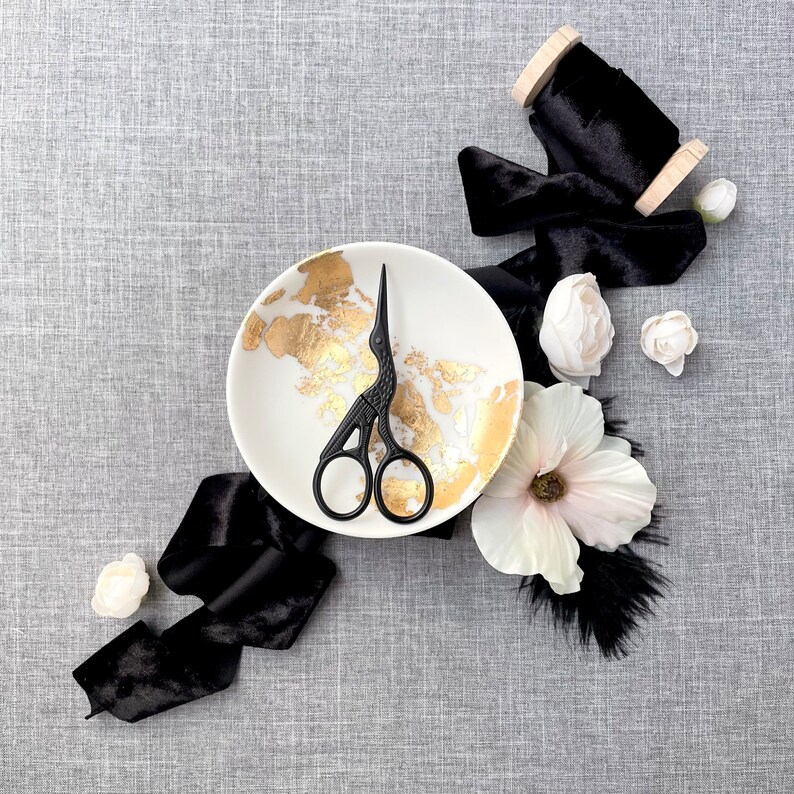 May include: A white ceramic dish with gold flecks, a black pair of small scissors, black velvet ribbon, and white flowers on a gray textured background.