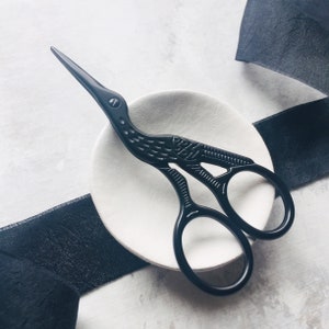 May include: Black decorative scissors with a bird design, resting on a white ceramic dish. The scissors have a detailed, intricate design and are a unique and stylish addition to any sewing or craft kit.