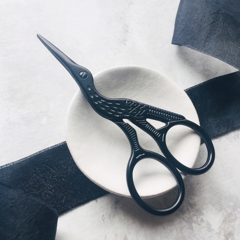 May include: Black decorative scissors with a bird design, resting on a white ceramic dish. The scissors have a detailed, intricate design and are a unique and stylish addition to any sewing or craft kit.