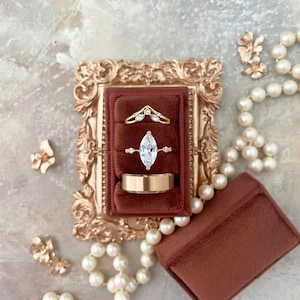 May include: A collection of gold rings displayed in a burgundy velvet ring box. The rings include a marquise-cut diamond ring, a curved band with small diamonds, and a simple gold band. The scene is complemented by pearl necklaces and floral accents.