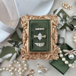 May include: An assortment of gold rings with various gemstone cuts, displayed in a green velvet-lined, ornate gold frame. The rings include baguette, marquise, and other diamond shapes. Pearls and floral accents complement the jewelry, suggesting a bridal or special occasion theme.