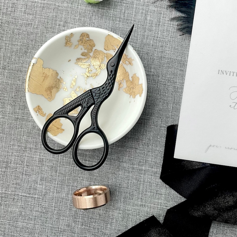 May include: A black bird-shaped scissor with gold accents sits in a white bowl with gold flecks. A gold ring is in the foreground.