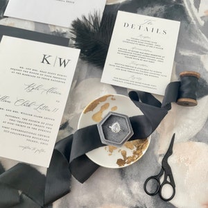 May include: A wedding invitation suite with black and white calligraphy on white card stock. The invitation features the couple's names and wedding date. The details card includes information about the ceremony and reception. A black ribbon and a small ring box with a diamond ring are also included in the image.