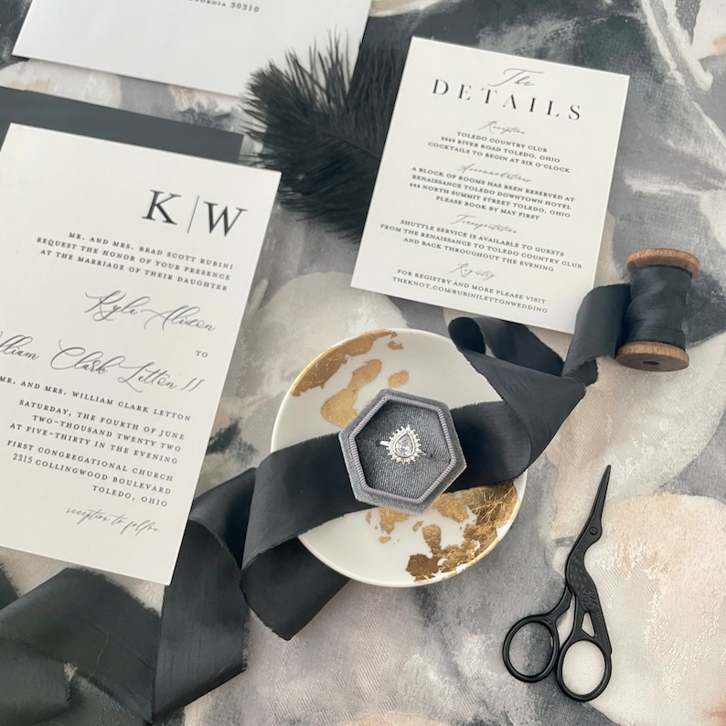 May include: A wedding invitation suite with black and white calligraphy on white card stock. The invitation features the couple's names and wedding date. The details card includes information about the ceremony and reception. A black ribbon and a small ring box with a diamond ring are also included in the image.