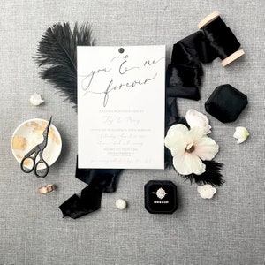 May include: A wedding invitation with black calligraphy that reads "you & me forever". The invitation is surrounded by black velvet ribbon, white flowers, a black feather, and a ring box.
