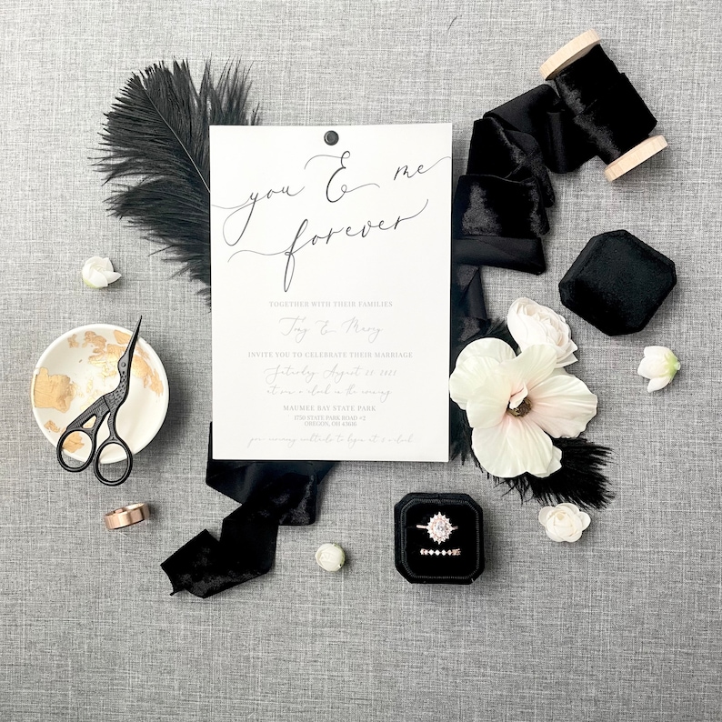 May include: A wedding invitation with black calligraphy that reads "you & me forever". The invitation is surrounded by black velvet ribbon, white flowers, a black feather, and a ring box.