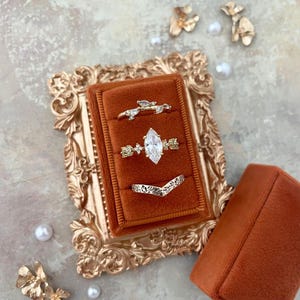 May include: Three gold rings with intricate designs are displayed in a rust-colored velvet ring box. The rings feature clear gemstones and delicate leaf and floral patterns. The box sits within an ornate gold frame, with decorative elements.
