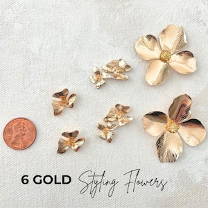 May include: Six gold-coloured styling flowers in various sizes are arranged on a white surface. The flowers have a detailed petal design and a textured finish. A penny is included for size reference. The text "6 GOLD Styling Flowers" is at the bottom.