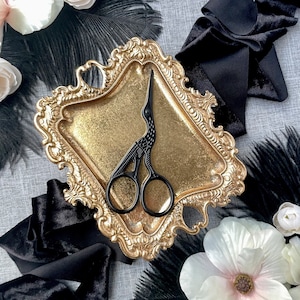 May include: A black ornate scissor with a bird design sits on a gold tray with a decorative border. The tray is surrounded by black feathers and fabric.