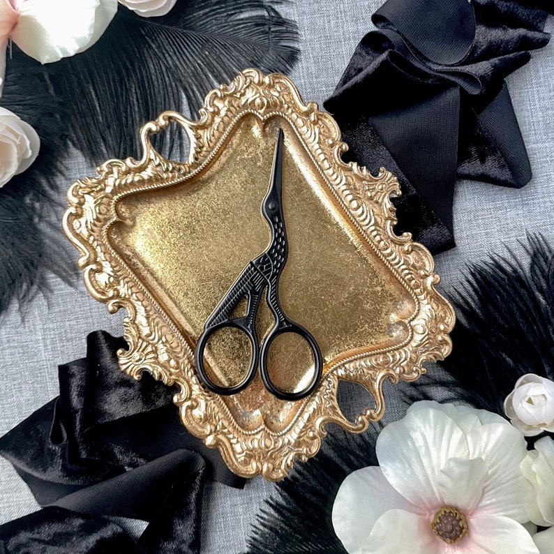 May include: A black ornate scissor with a bird design sits on a gold tray with a decorative border. The tray is surrounded by black feathers and fabric.