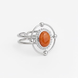 Può includere: Anello color argento con una gemma ovale marrone-arancio al centro. L'anello ha un design a doppia fascia con una cornice circolare attorno alla gemma, accentuata da piccole perline e sfere argentate.