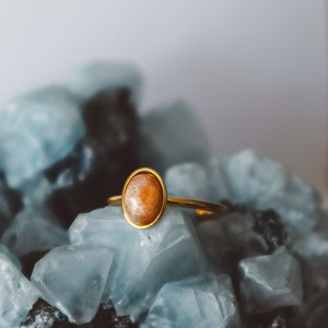 May include: A gold ring featuring a light brown oval gemstone set in a bezel setting. The ring is resting on a bed of blue and grey crystals.