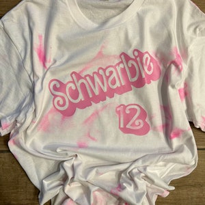 May include: White t-shirt with a pink tie-dye pattern. The shirt has a pink graphic that says "Schwarbie" with the number "12" underneath.