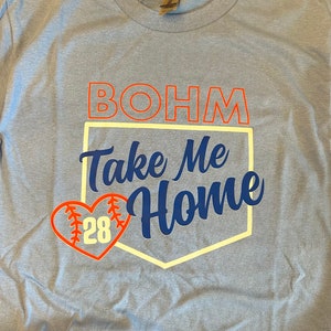 May include: Light blue t-shirt with a white baseball diamond graphic. The graphic has the text "BOHM Take Me Home" in blue and orange. A red heart with a baseball graphic and the number "28" is in the corner of the diamond.
