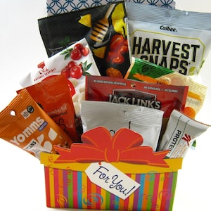 May include: A colorful gift basket filled with various snack packages, including "Harvest Snaps" and "Jack Link's." The basket has a red bow and a tag that says "For You!"