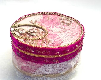 Op de afbeelding: Een ovale sieradendoos met een roze en wit bloemen kanten ontwerp. De doos heeft een fuchsia fluwelen band, gouden afwerking en een decoratieve gouden ketting. Een kleine, heldere edelsteen is bevestigd aan de bovenkant. Het is een decoratief item.