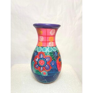 May include: A ceramic vase featuring a purple rim and a pink upper section decorated with small yellow and white flower motifs. The lower body displays large, vibrant red and blue floral patterns against a dark blue base with green leaves.
