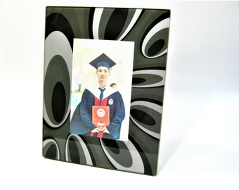 May include: A black and silver photo frame with a swirling pattern. The frame has a rectangular opening for a photo. A photo of a person in a graduation gown and cap is visible in the opening.