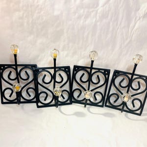 May include: Four black metal wall hooks with a decorative heart design. Each hook has a clear, faceted knob at the top and bottom, and a small, yellow-toned accent. The hooks are square and have a scrolled design.