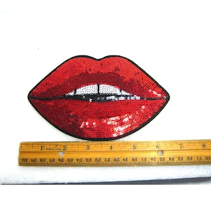May include: A red sequin lip-shaped patch with a black border and white teeth detail, positioned above a wooden ruler that shows the patch is approximately 8 inches wide. The sequins create a reflective surface across the lips.