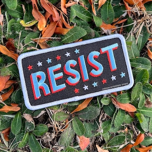 May include: A black fabric patch with white stitching around the edges. The patch features the word "RESIST" in blue and red letters with white stars around the text. The text "HANNAH ROSENGREN STUDIO" is printed in white in the bottom right corner of the patch.