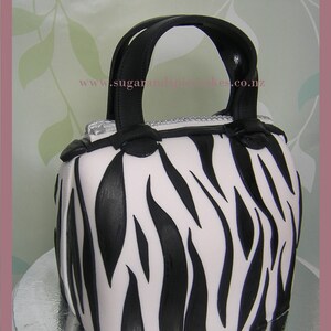 May include: A black and white zebra-print handbag cake with black handles. The cake is decorated with a silver zipper detail and the website www.sugarandspice.co.nz.