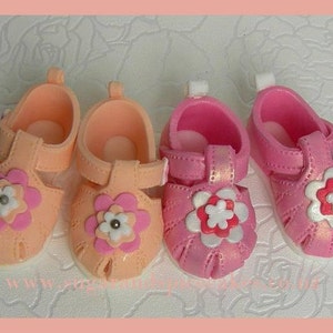 May include: Four pairs of baby sandals in shades of pink and peach. Each sandal features a flower applique with white and pink petals and a silver center. The sandals have a strap closure and are designed for infants.