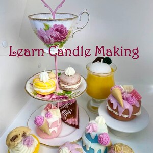 Könnte beinhalten: Ein mehrstöckiger Kuchenständer mit einem rosa Griff und floralem Design hält verschiedene duftende Kerzen in Form von Desserts, darunter Macarons, Cupcakes und ein Stück Kuchen. Der Text "Learn Candle Making" ist im Bild sichtbar.