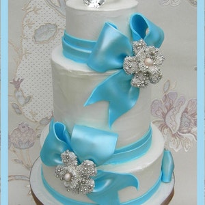 May include: A three-tiered white wedding cake decorated with blue satin ribbon bows and silver and pearl embellishments. A large diamond ring sits on top of the cake.