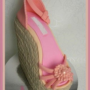 May include: A pink and beige wedge shoe cake with a flower detail. The cake is decorated to look like a real shoe.