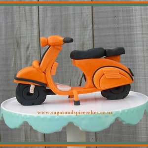 May include: Orange and black fondant scooter cake decoration. The scooter is on a white cake stand with a light blue base.
