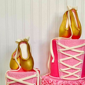 May include: Two pink and white tiered cakes decorated with gold ballet shoes. The top tier has a pink corset design with white laces. The bottom tier has a pink ruffle design.