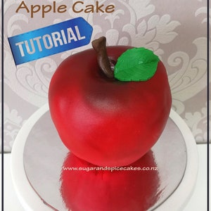Peut inclure: Un gâteau en forme de pomme rouge avec une feuille verte sur le dessus. Le gâteau est sur un support à gâteau blanc. Le texte "Teacher's Pet Apple Cake TUTORIAL" est visible sur l'image.