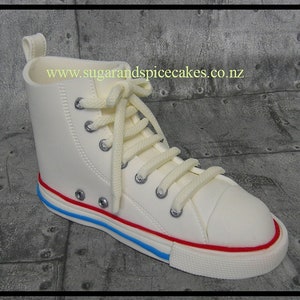 Peut inclure: Des baskets blanches montantes avec des lacets blancs et une bande rouge et bleue autour de la semelle. Les baskets sont en pâte à sucre et sont un gâteau.