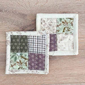 May include: Two square fabric coasters with a patchwork design. Each coaster features a cream-colored border and a central pattern of geometric shapes, floral prints, and a gingham pattern in shades of green, brown, and purple. The coasters are placed on a wooden surface.