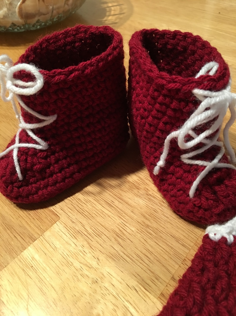 Puede incluir: Un par de botines de beb&eacute; hechos a mano, tejidos a ganchillo, de color rojo intenso. Los botines tienen cordones blancos y un patr&oacute;n de puntada texturizada. Los botines est&aacute;n dise&ntilde;ados para parecer botas en miniatura.