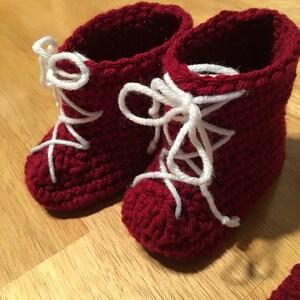 Puede incluir: Un par de patucos de beb&eacute; de ganchillo rojo hechos a mano con cordones blancos. Los patucos est&aacute;n dise&ntilde;ados para parecer botas en miniatura, con un patr&oacute;n de puntada texturizada y un dise&ntilde;o cl&aacute;sico de cordones. Se muestran sobre una superficie de madera.