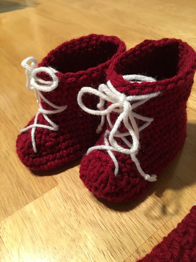 Puede incluir: Un par de patucos de beb&eacute; de ganchillo rojo hechos a mano con cordones blancos. Los patucos est&aacute;n dise&ntilde;ados para parecer botas en miniatura, con un patr&oacute;n de puntada texturizada y un dise&ntilde;o cl&aacute;sico de cordones. Se muestran sobre una superficie de madera.