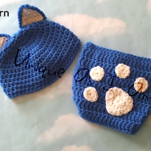 May include: A blue crocheted hat with cat ears and a matching blue crocheted diaper with white paw prints. The pattern is labeled "Pattern".